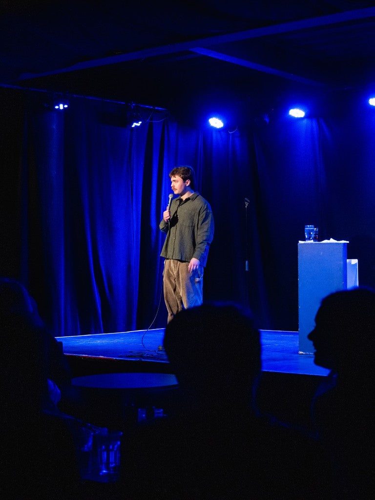 Comedian performing on a blue-lit stage during Belfast Whiskey Week fringe programming