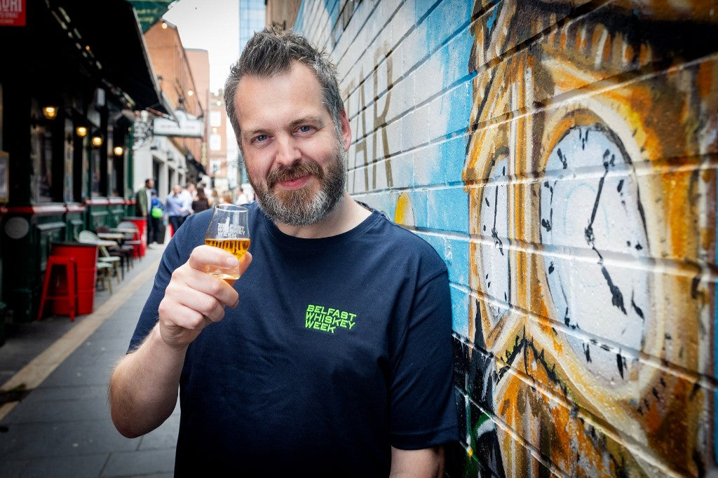 Festival director Paul Kane in a Belfast alleyway holding a whiskey tasting glass, wearing a Belfast Whiskey Week t-shirt