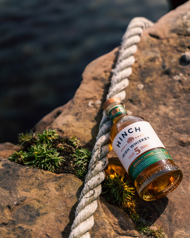 Bottle of Hinch 5 Year Old Double Wood Irish whiskey resting on coastal rocks by a rope